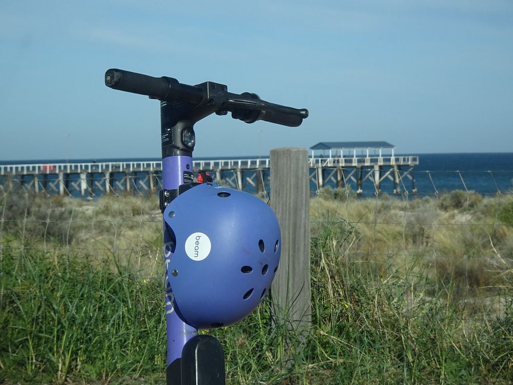An e-scooter parked by the beach with a helmet hanging from its handlebars